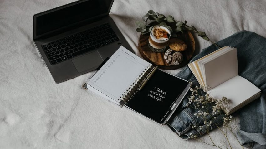 A cozy flat lay of a laptop, journals, and snacks on a bed for a relaxed work-from-home setup.
